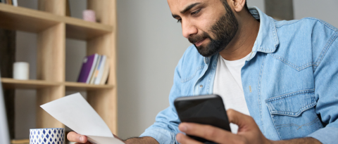 Young businessman holding phone reading bank receipt calculating taxe